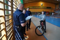 Policjanci na sali gimnastycznej rozmawiają z uczniami, po lewej stronie uczeń siedzi na rowerze