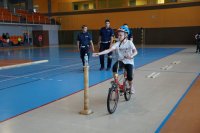 Na pierwszym planie uczennica na rowerze jedzie na torze przeszkód, na drugim planie dwóch policjantów